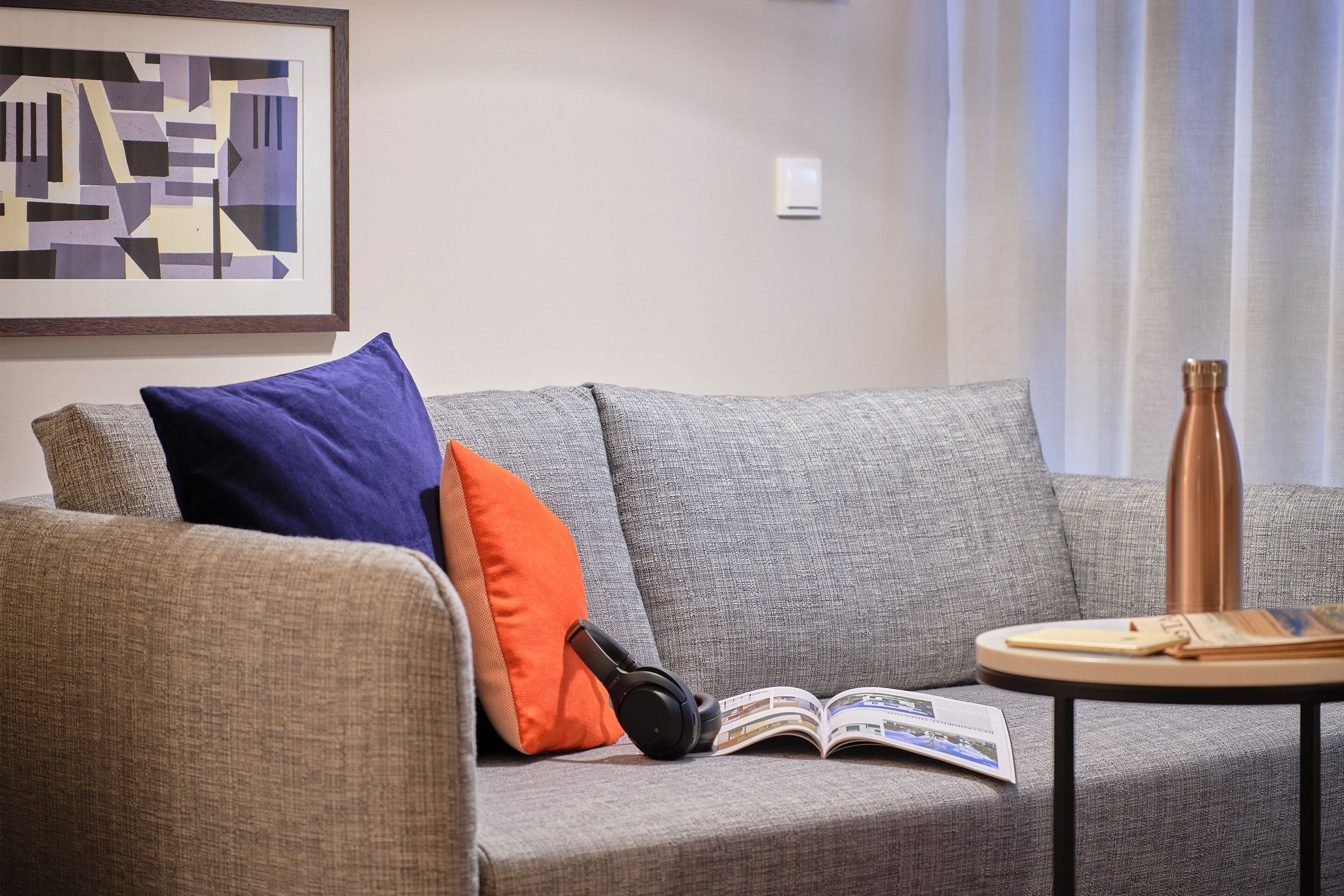 Un canapé gris avec un coussin bleu et orange, une paire d’écouteurs et un magazine ouvert sur une petite table ronde.