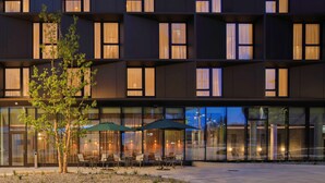 Un edificio moderno con varios pisos, ventanas amplias y mesas exteriores con sombrillas.