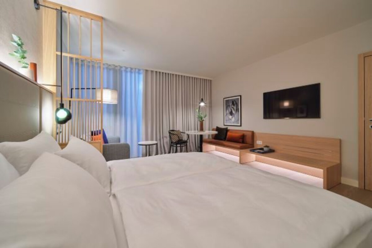 Una habitación de hotel moderna con una cama grande, un escritorio, una silla, un sofá, un televisor y una ventana con cortinas.