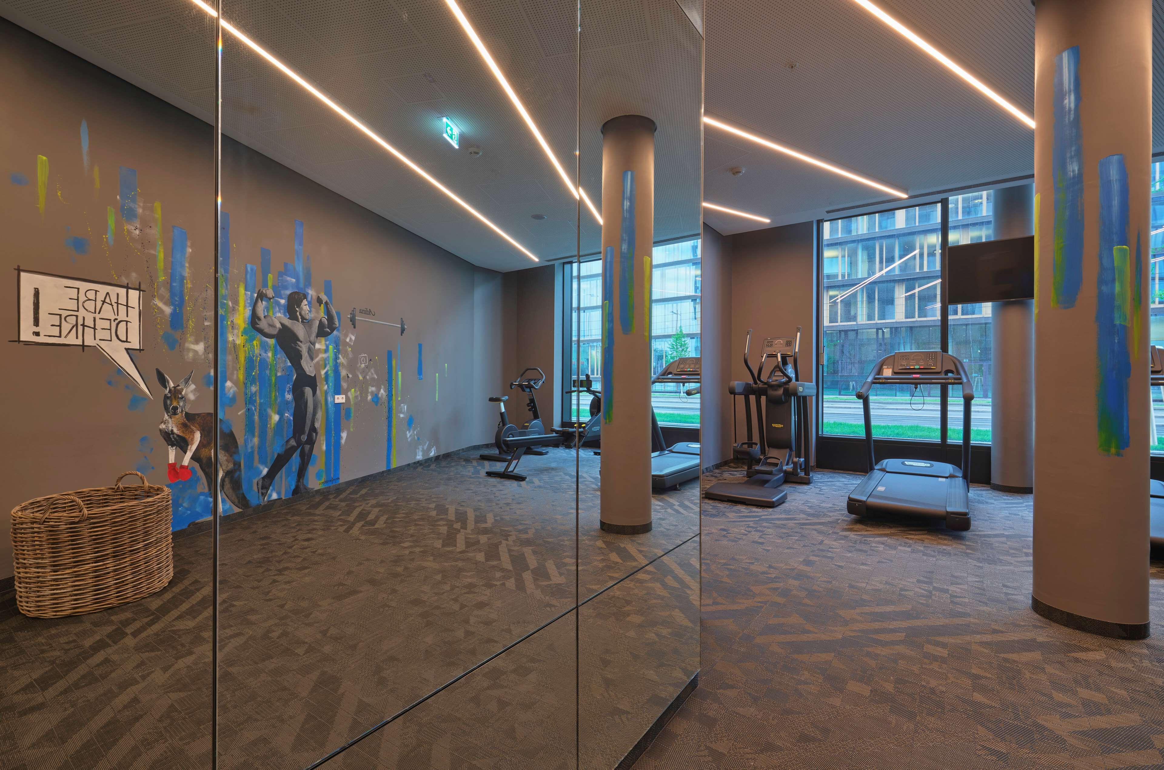 A modern gym with exercise equipment, large windows, and a mural on the wall.