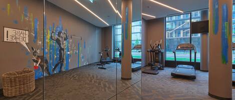 A modern gym with exercise equipment, large windows, and a mural on the wall.