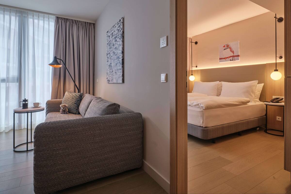 Una habitación de hotel moderna con un sofá, una mesita con una cafetera, una cama con ropa de cama blanca y una obra de arte colgada en la pared.