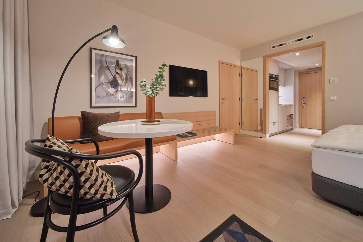 Una habitación de hotel moderna con una mesa redonda, un sofá, una silla, una cama, un televisor y una puerta que da a una cocina.