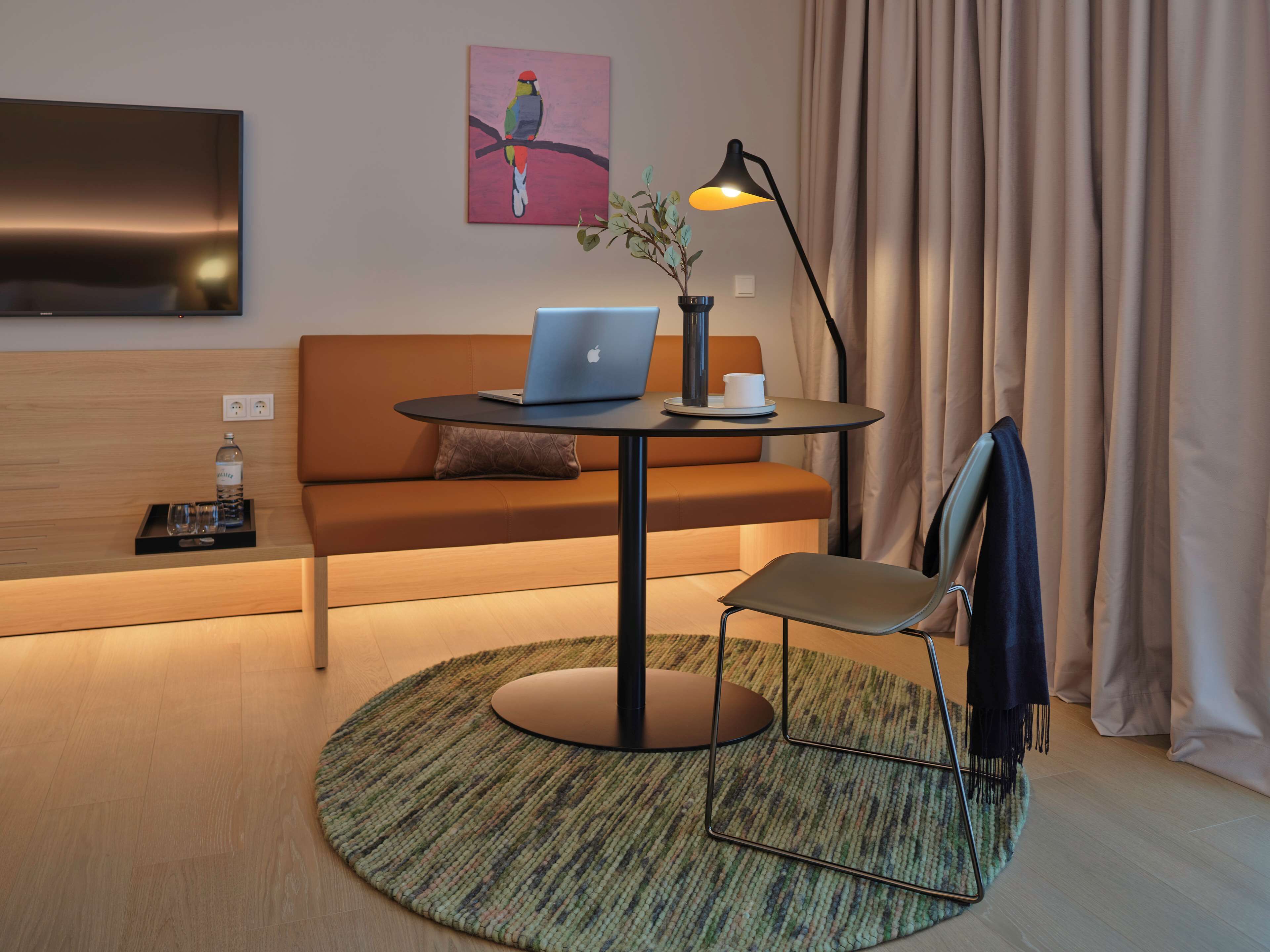 Ein modernes Hotelzimmer mit einer Couch, einem runden Tisch mit einem Laptop, einer Lampe und einem kleinen Topfpflanzen.