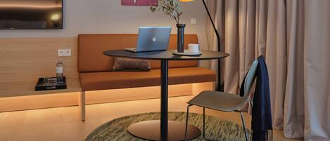 A modern hotel room with a sofa, a round table with a laptop, a lamp, and a small potted plant.