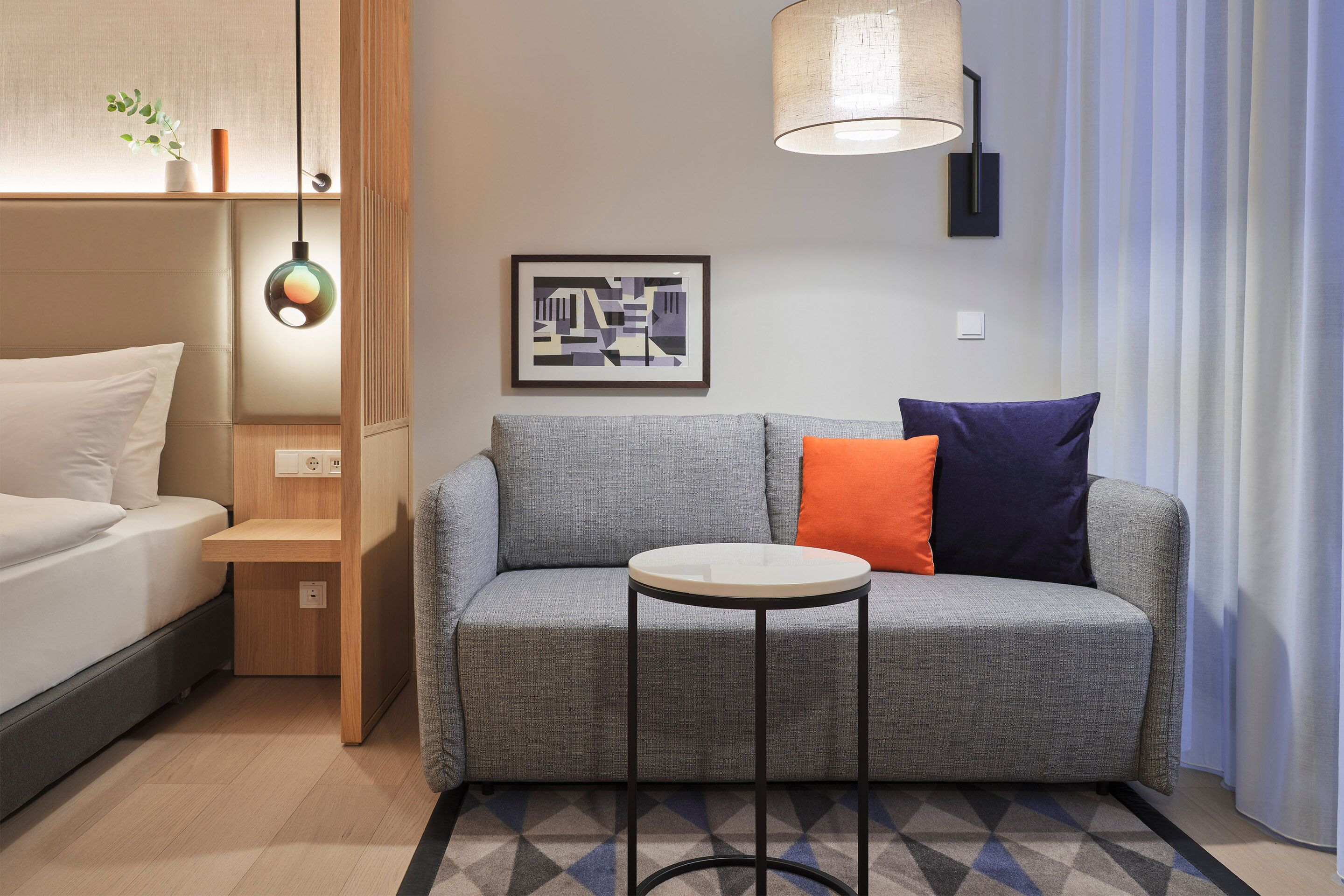 Ein modernes Hotelzimmer mit grauem Sofa, einem kleinen runden Tisch, einem Bett mit weißer Bettwäsche, einer Wandlampe und einem gerahmten Bild.