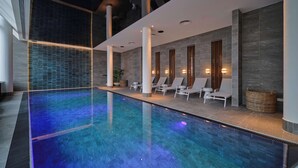 Un área de piscina interior moderna con una piscina azul cristalina, sillas de descanso e iluminación en la pared.