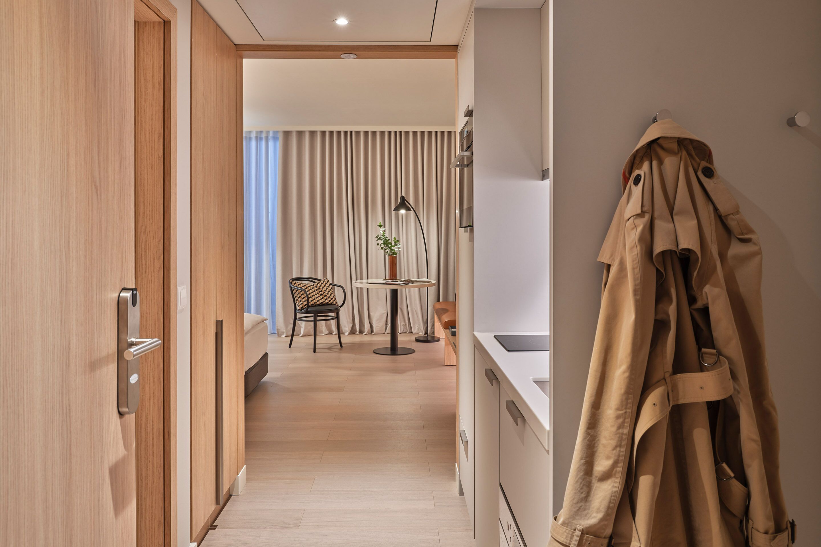 A hotel room with a wooden door, a coat rack, a dining area with a table and chairs, and a view of the room through a curtain.