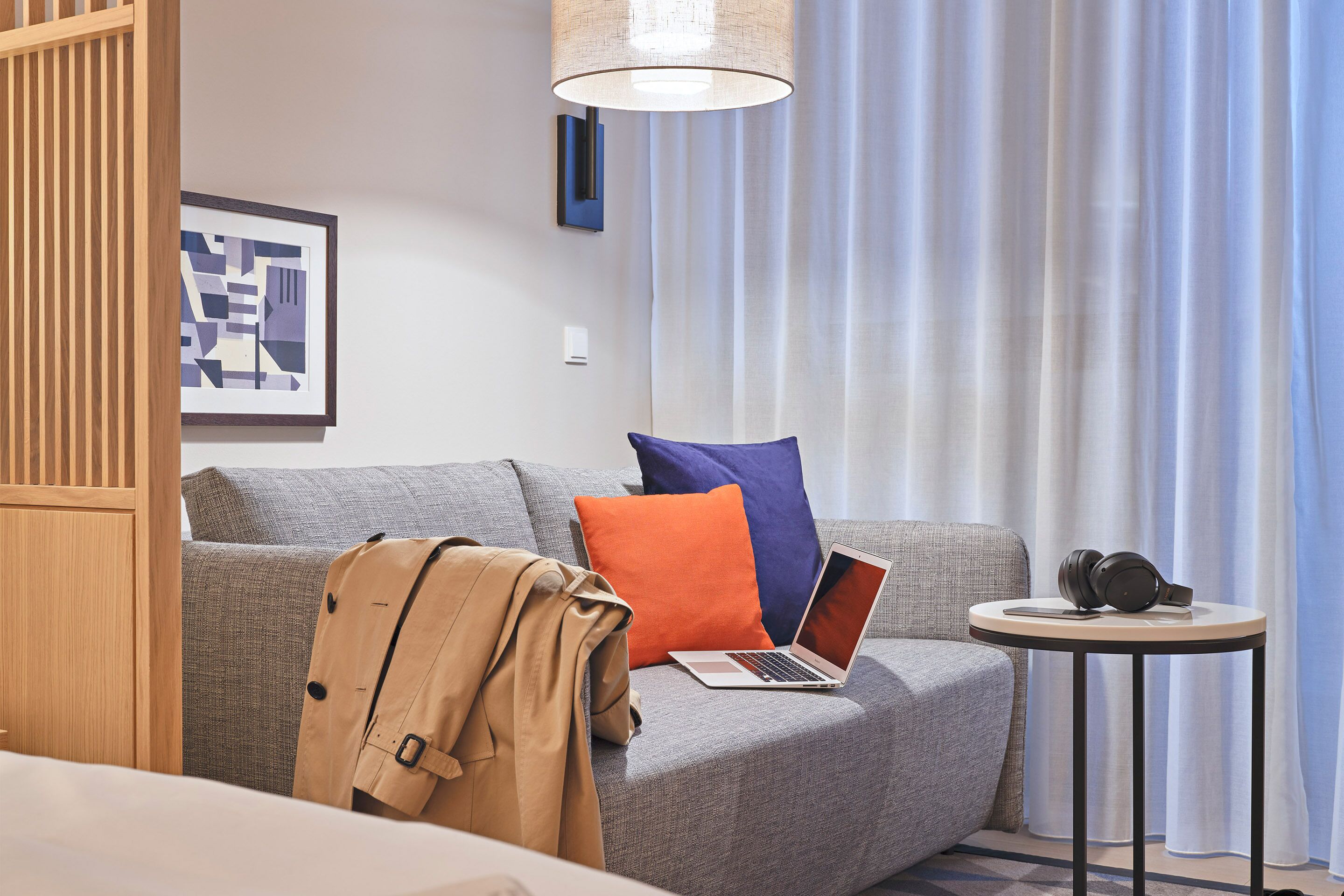 Quarto de hotel moderno com sofá cinza, um laptop, auscultadores e uma mesa redonda pequena.
