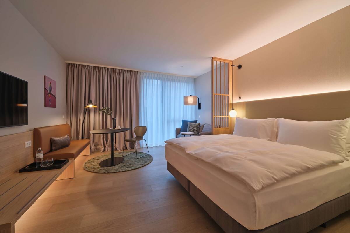 Una habitación de hotel moderna con una cama grande, un sofá, una mesita y un televisor.