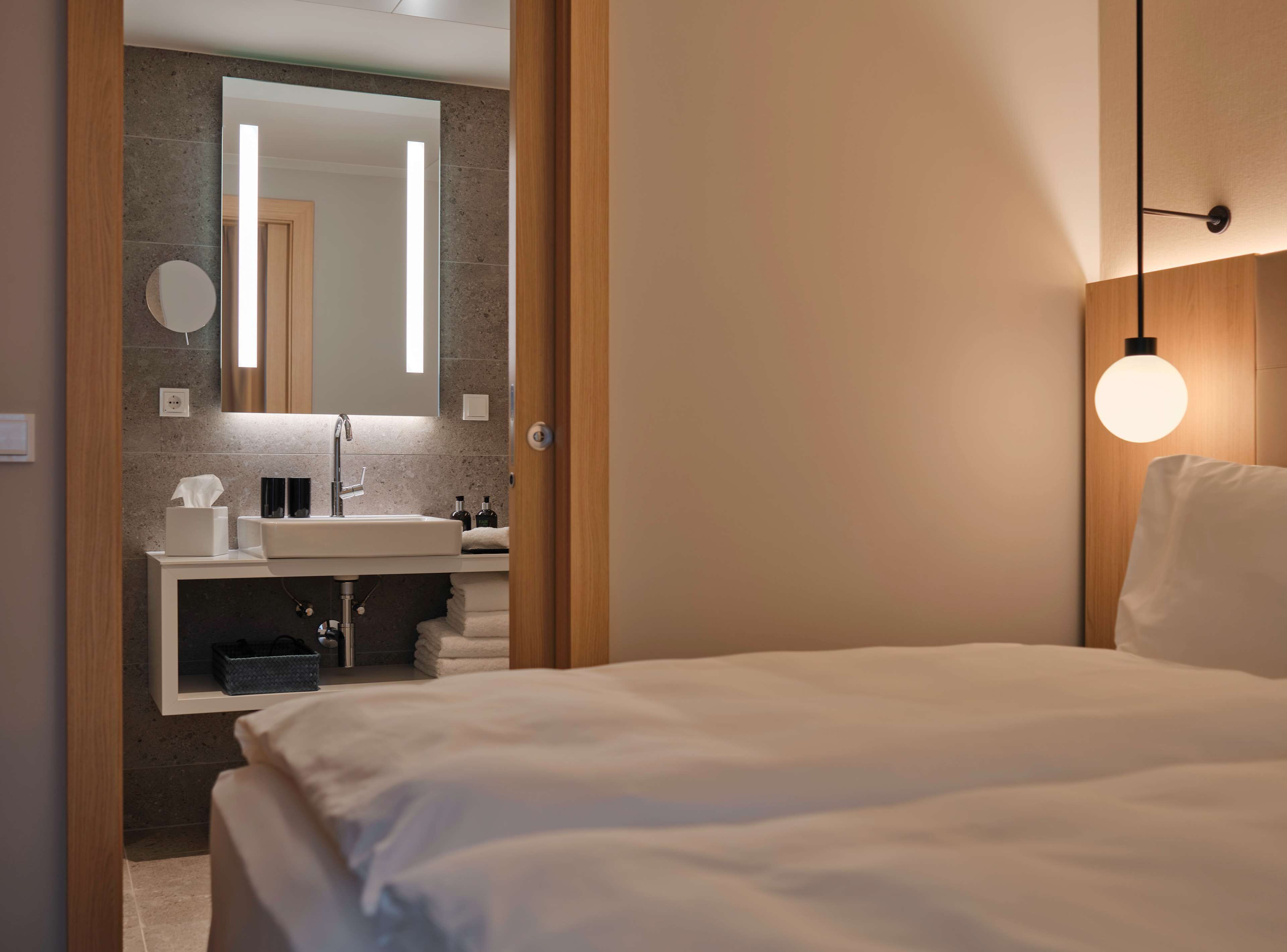 Ein modernes Hotelzimmer mit einem großen Bett, einem Badezimmer mit Waschbecken und Spiegel sowie einer Pendelleuchte.