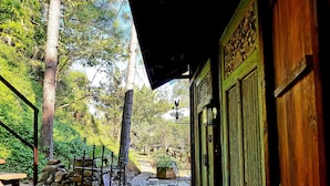 Un sendero de piedra que conduce a un edificio de madera con una puerta verde, rodeado de abundante vegetación y un muro de piedra.