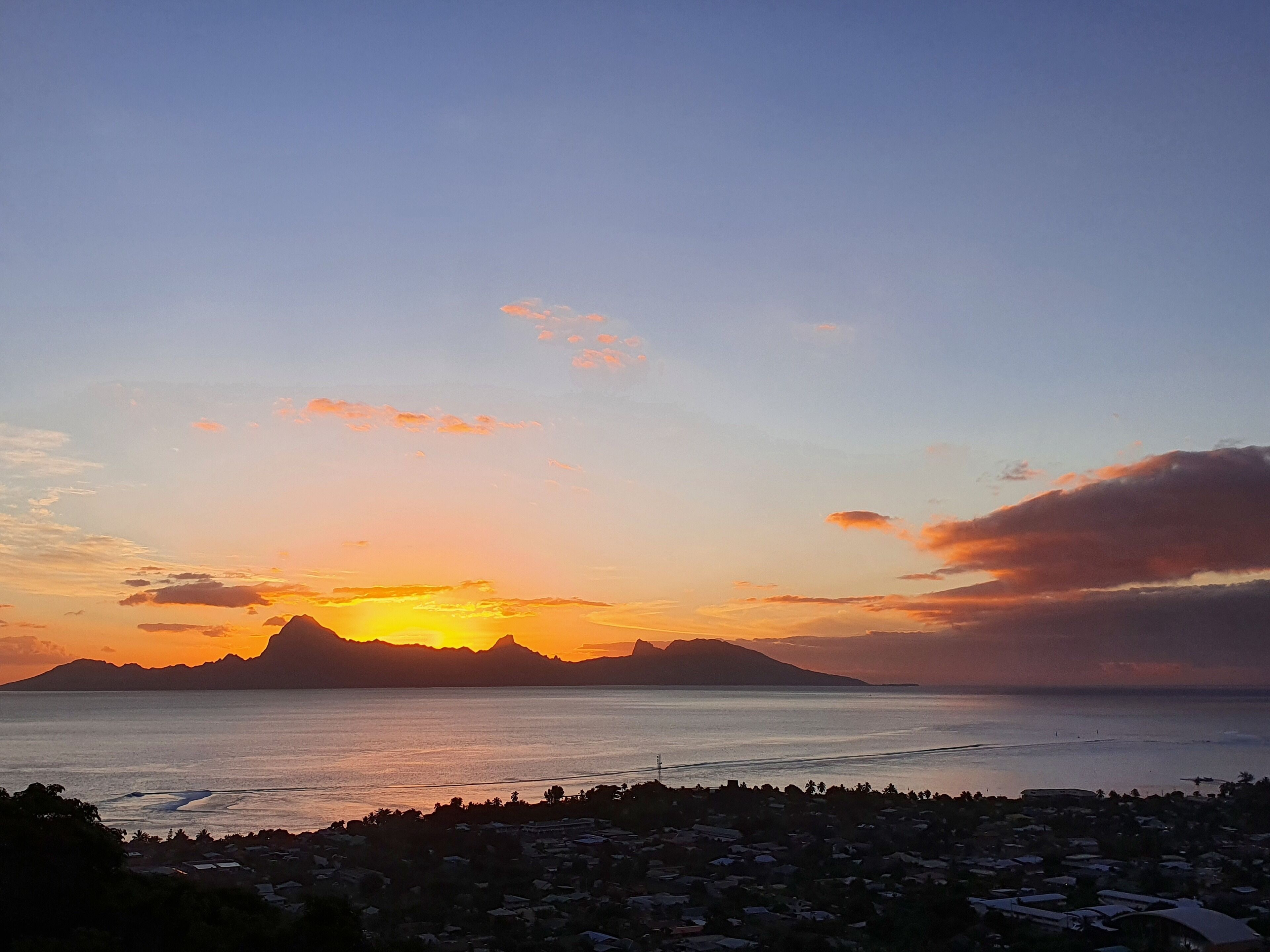 A coastal landscape with a sunset over the sea, mountains in the background, and a residential area in the foreground.