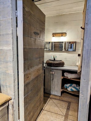 Une salle de bain rustique avec une porte en bois, un lavabo doté d’un bac rond, un miroir encadré d’un motif géométrique et un porte-serviettes.