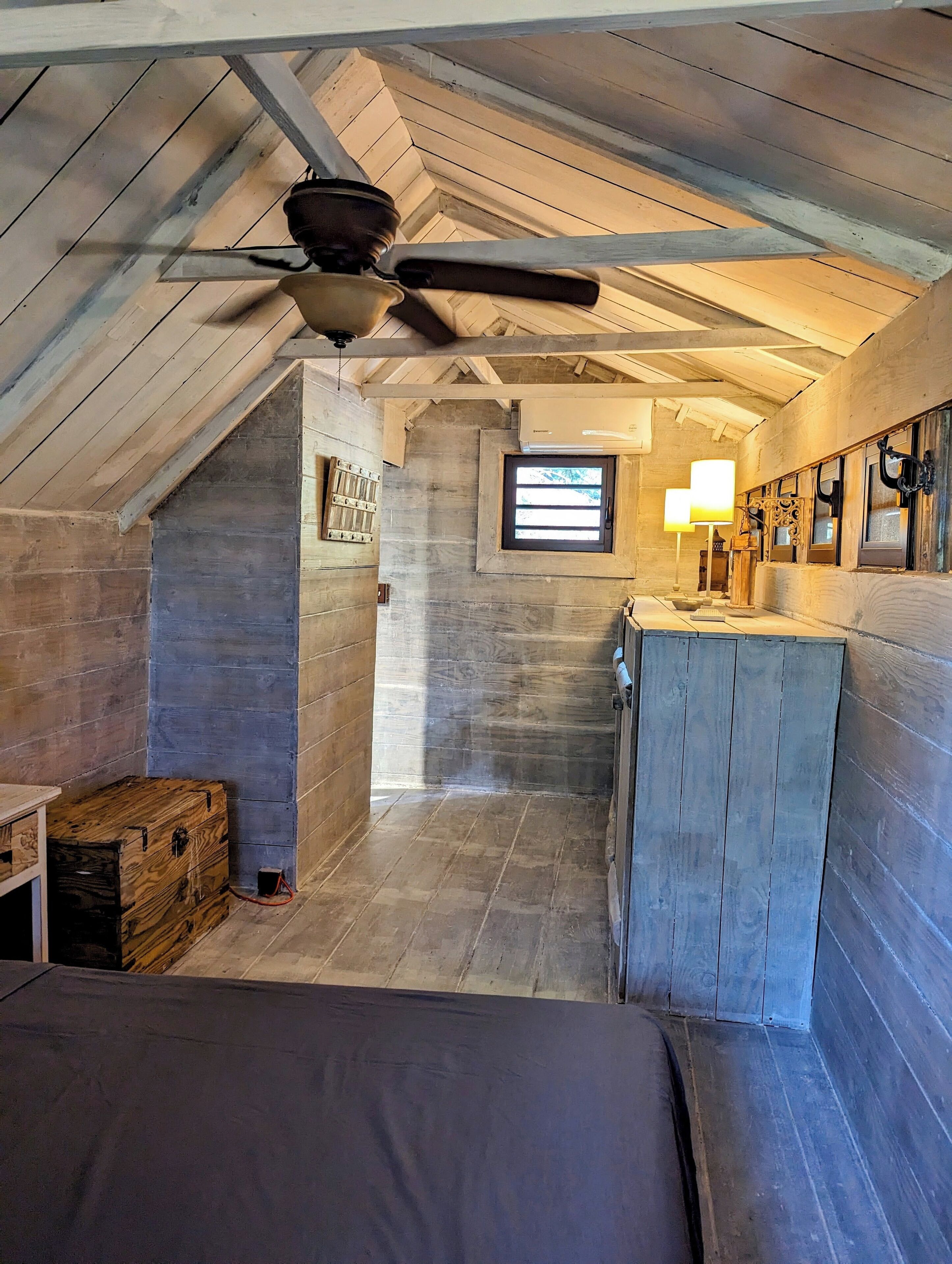 Une chambre mansardée au style rustique, avec un plafond en pente, un ventilateur de plafond, une petite fenêtre et un banc en bois.