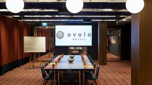 Een vergaderruimte met een groot scherm waarop het logo van Ovolo Hotels staat, een lange tafel met stoelen en een vloerbedekking.