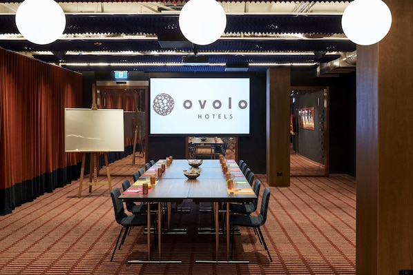 Et mødelokale med et stort skærmbillede der viser Ovolo Hotels' logo, et langt bord med stole, og et tæppe på gulvet.