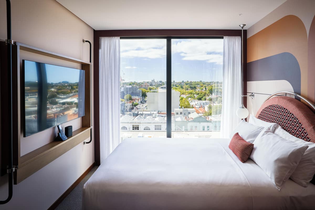 Una habitación de hotel con una cama grande, un televisor de pantalla plana montado en la pared, un ventanal con vistas a la ciudad y un cabecero moderno.