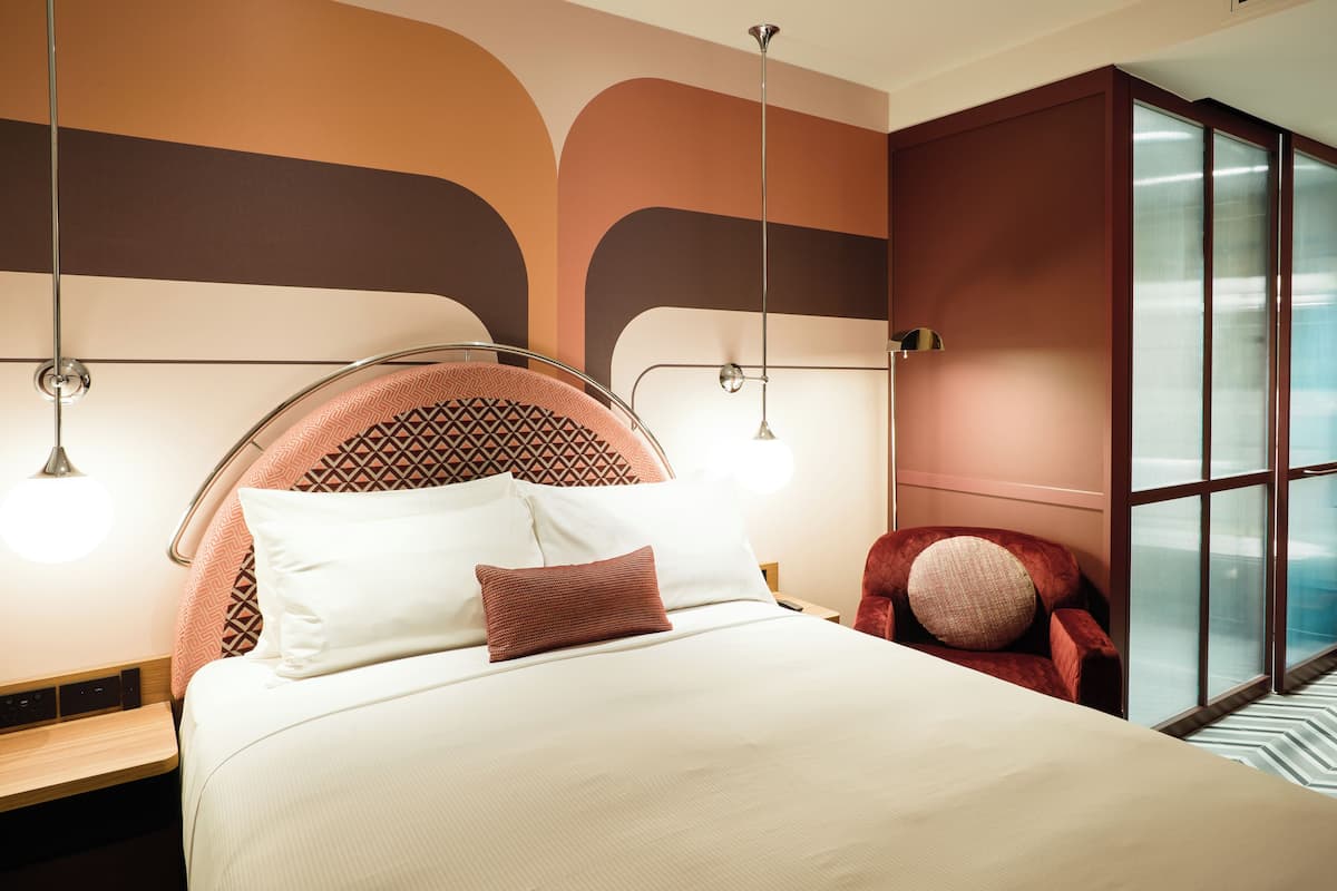 Una habitación de hotel moderna con una cama, mesitas de noche, un sillón rojo y un ventanal grande.
