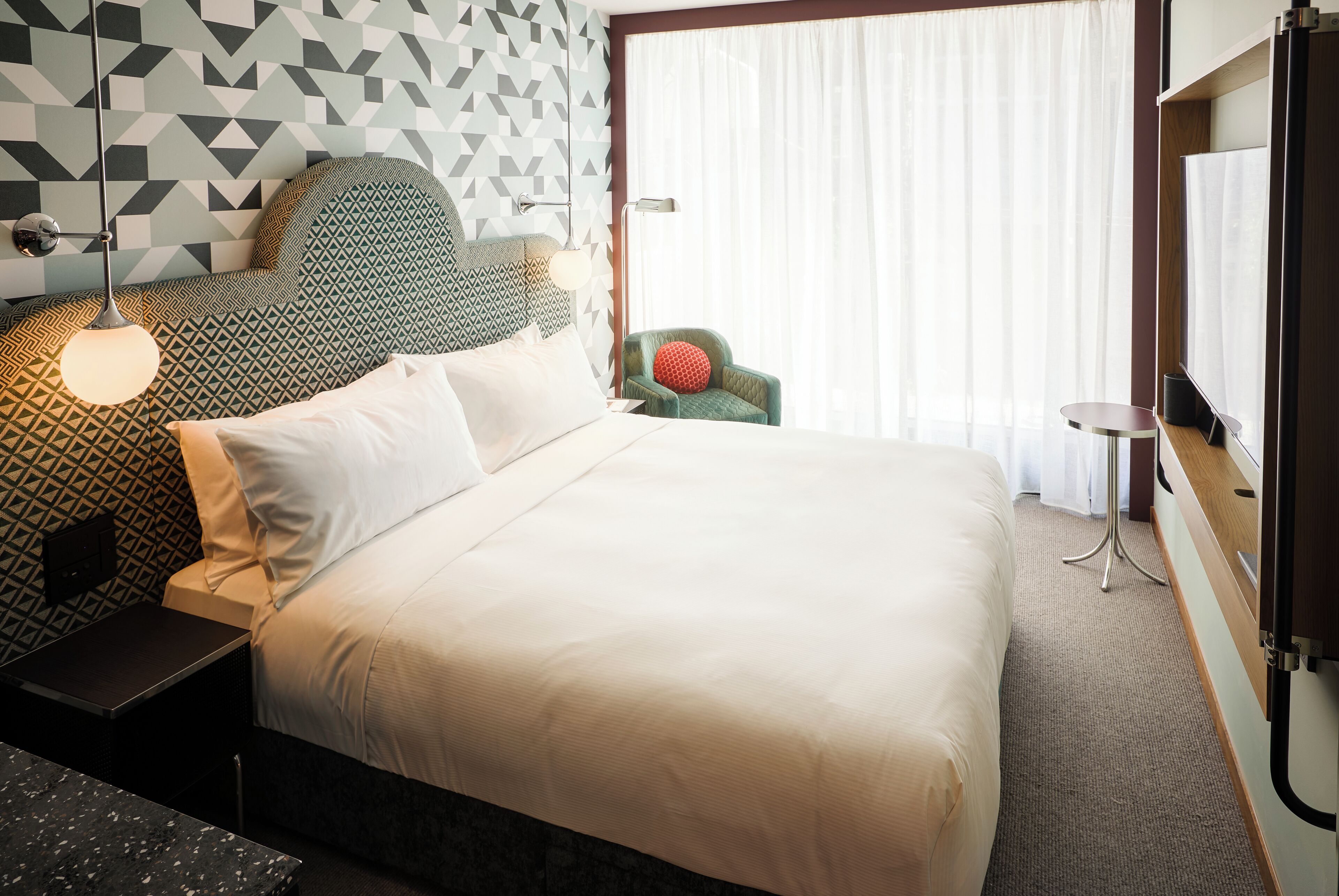 A neatly made bed with white linens, a geometric patterned headboard, a green armchair with a red cushion, a small round table, and a large window with sheer curtains.