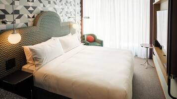 A neatly made bed with white linens, a geometric patterned headboard, a green armchair with a red cushion, a small round table, and a large window with sheer curtains.