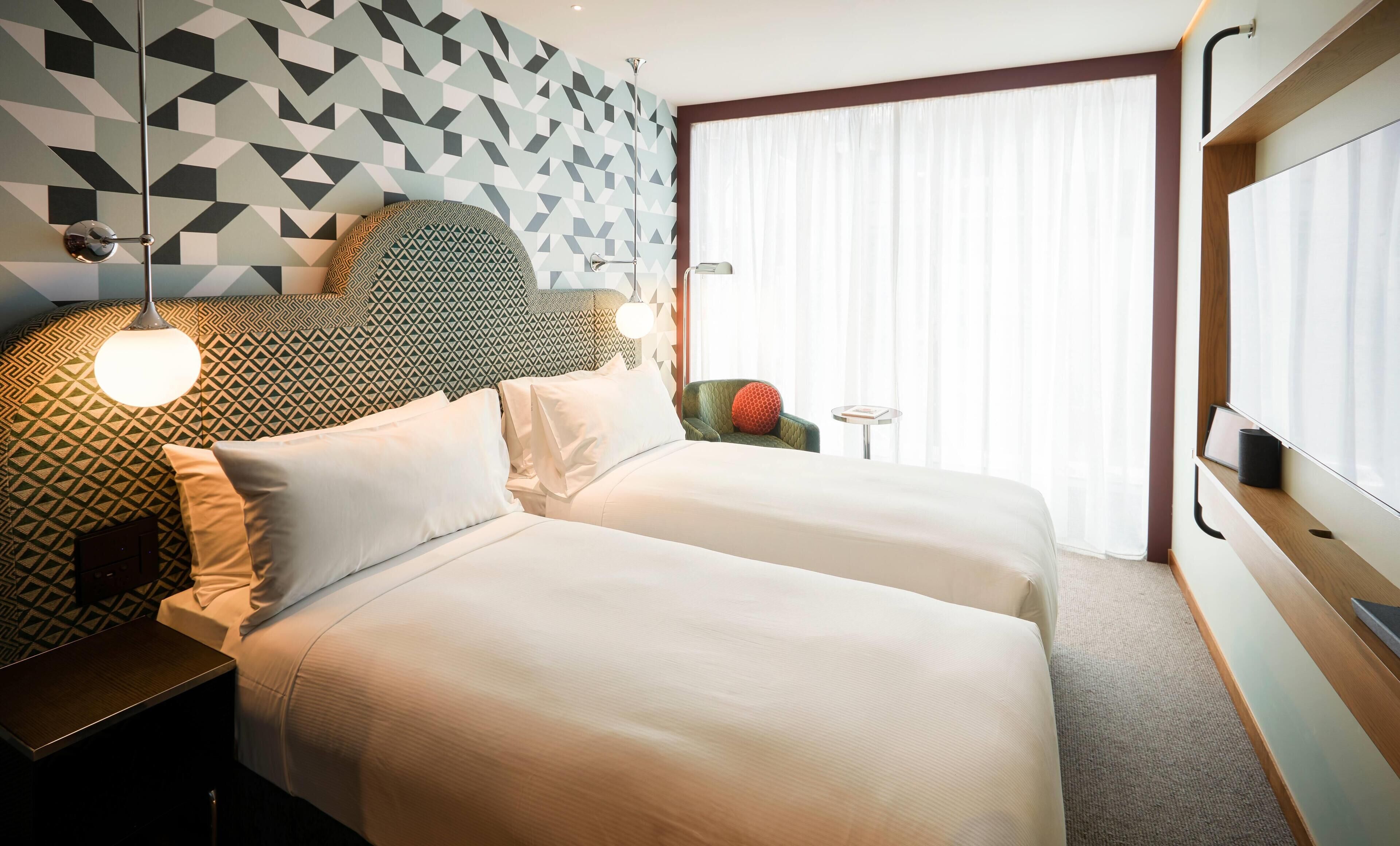Habitación de hotel con dos camas, un papel tapiz de diseño geométrico, una ventana con cortinas traslúcidas y una mesita de noche con lámpara.