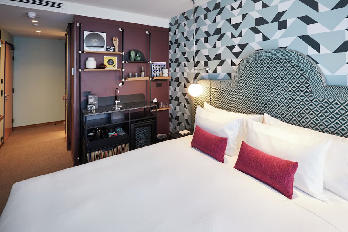 Habitación de hotel con cama, cabecera de diseño geométrico, repisa con varios objetos y pared con motivo geométrico.