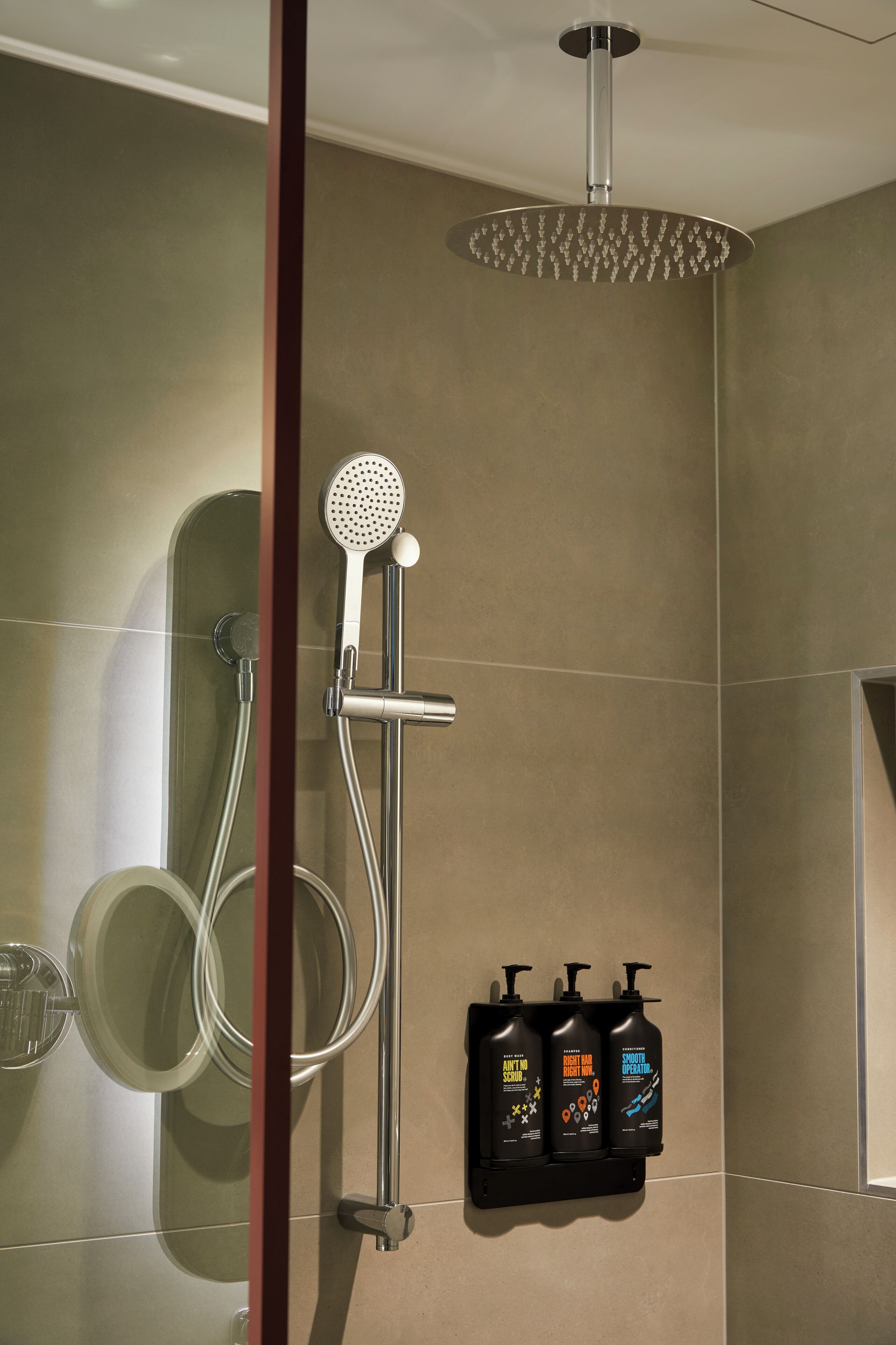 Eine Dusche mit Handbrause und Wandarmatur, ein gläsernes Duschgehäuse und drei Pflegeflaschen auf einem Wandregal.