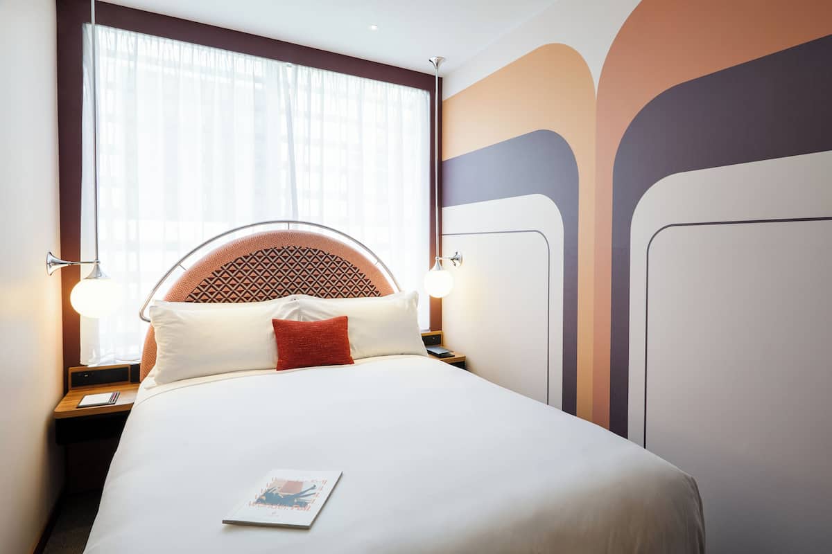 Una cama bien hecha con un cabecero estampado, lámparas de noche y una ventana con cortinas traslúcidas.