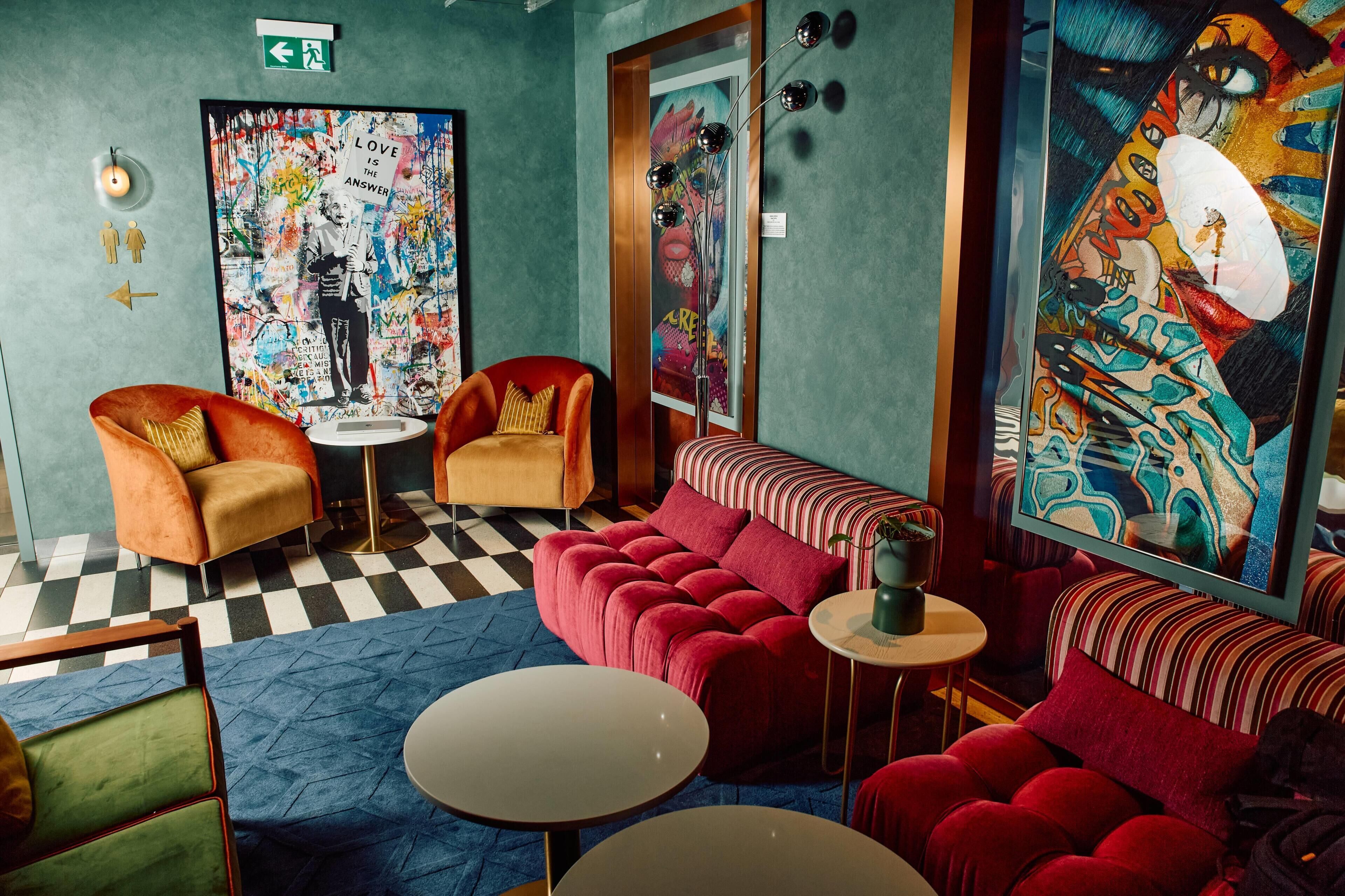 A room with a checkered floor, a red sofa, a small round table, and a chair with a cushion. There are two paintings on the walls, one with a figure and text, and another with a colorful abstract design.