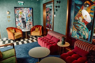 A room with a checkered floor, a red sofa, a small round table, and a chair with a cushion. There are two paintings on the walls, one with a figure and text, and another with a colorful abstract design.