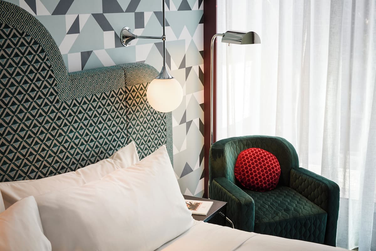 Habitación de hotel con cama, sillón verde, almohada roja, pared con estampado geométrico y lámpara colgante.