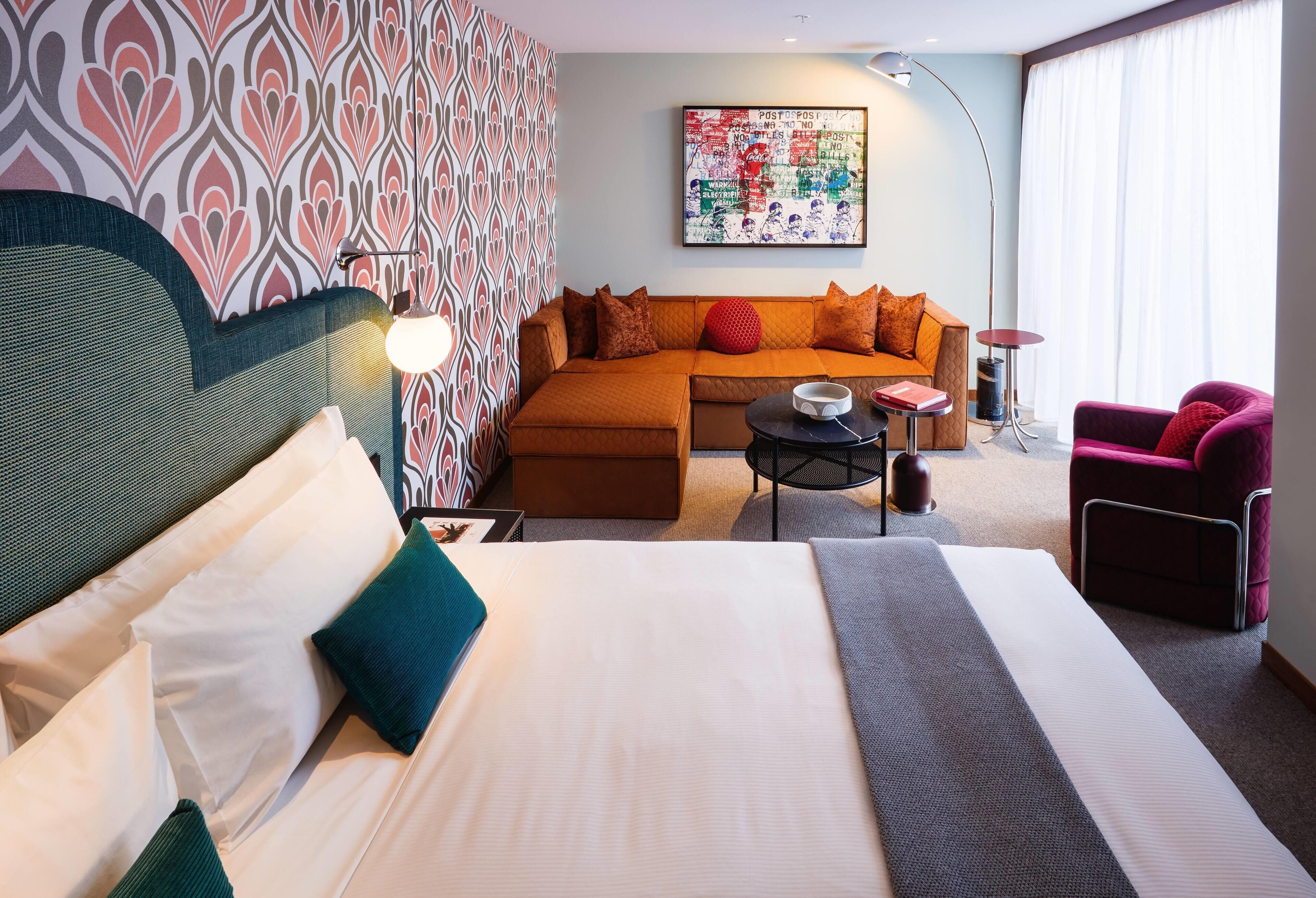 A hotel room with a large bed, a sofa, a small table, and a chair. There is a wall with a patterned wallpaper and a painting above the sofa.