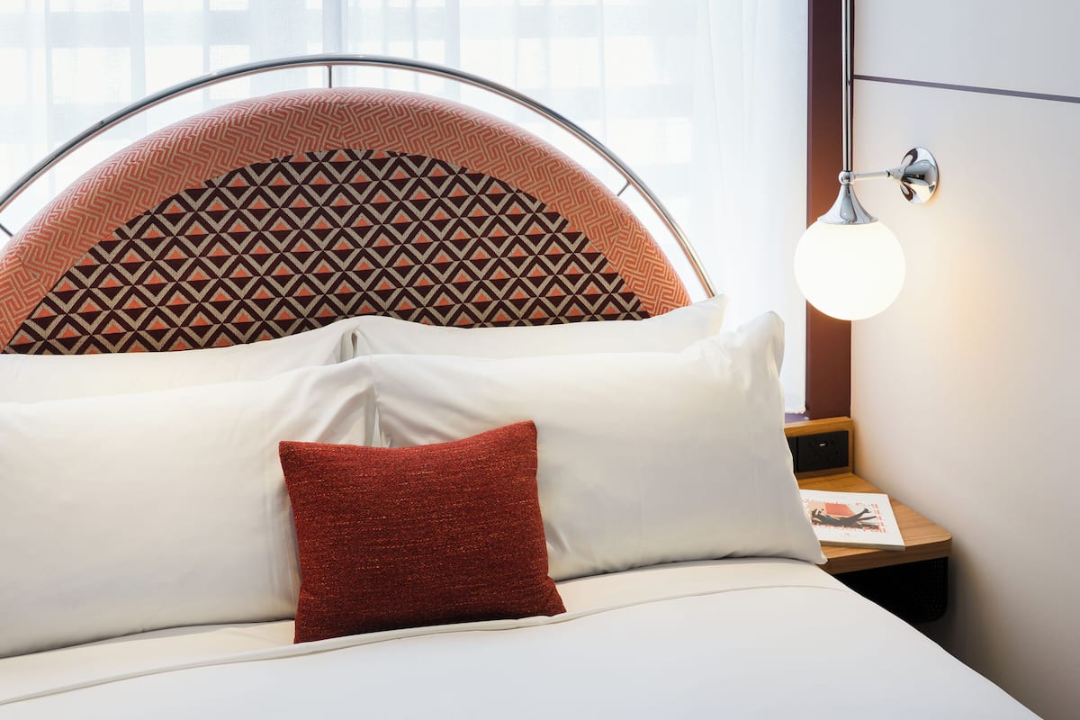 Una cama con cabecera, almohadas y una mesita de noche con lámpara.