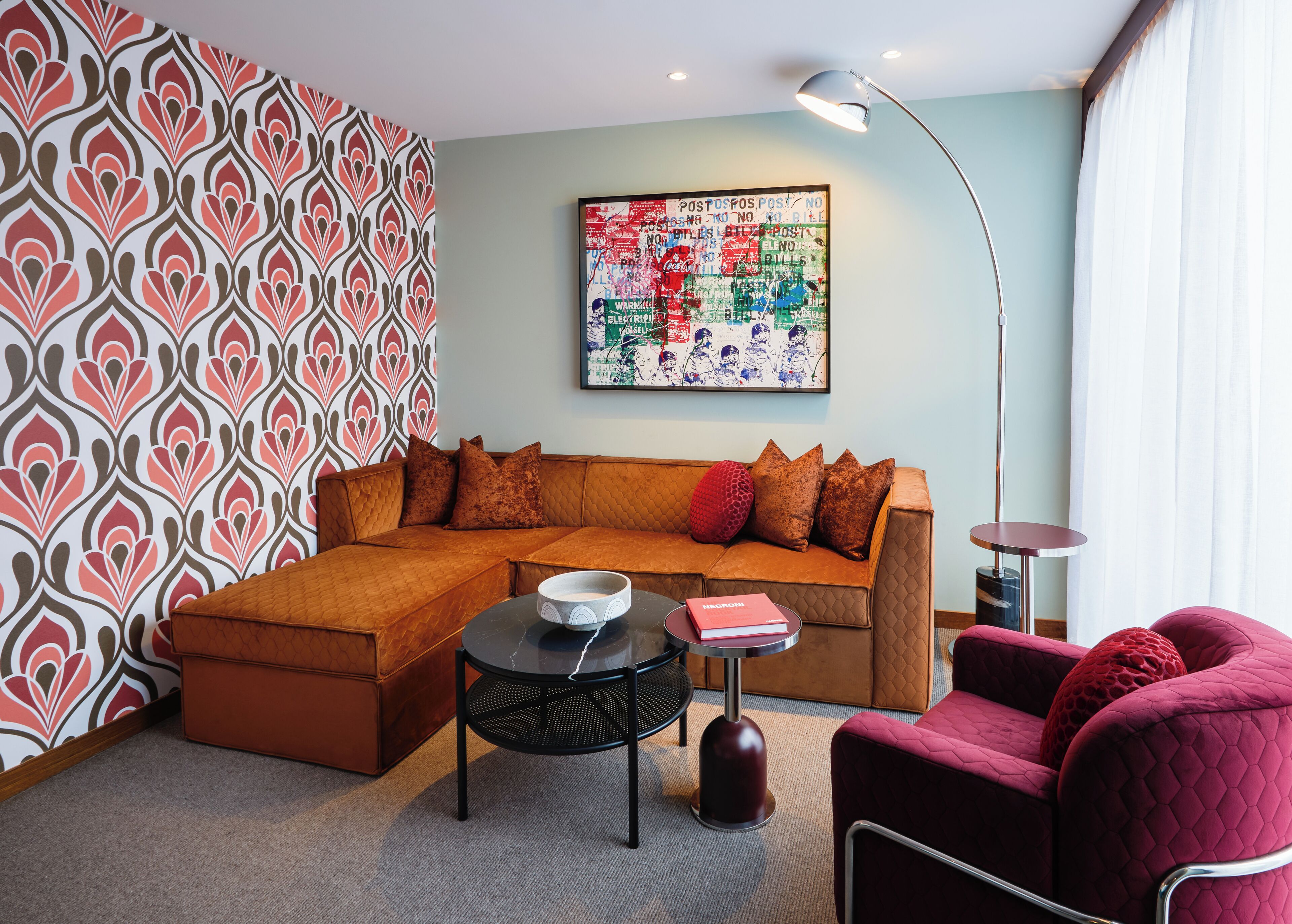 A living room with a sectional sofa, a red armchair, a round table with a book, and a colorful abstract painting on the wall.