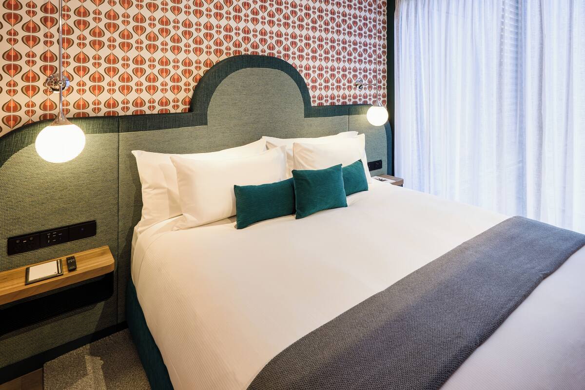 Una cama bien hecha con almohadas blancas y turquesa, cabecera con diseño geométrico, mesita de noche con lámpara y pared con estampado.