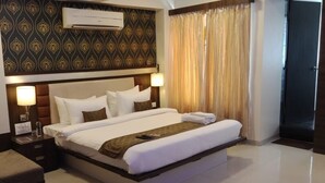 A hotel room with a large bed, a bedside table with a lamp, a wall-mounted air conditioner, and a curtain on the window. - Hotel Sai Vijay (Nashik)