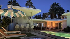 Un área de piscina moderna al aire libre con chimenea, sillas de descanso y zona de bar.