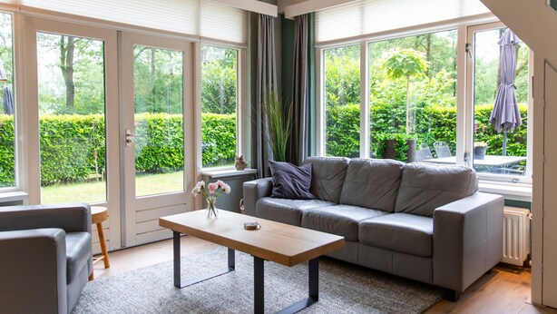 Un salon moderne avec un canapé gris, une table basse en bois et un fauteuil gris. De grandes fenêtres sont ornées de rideaux, et l’on peut admirer la verdure à l’extérieur. Un radiateur est installé au sol.