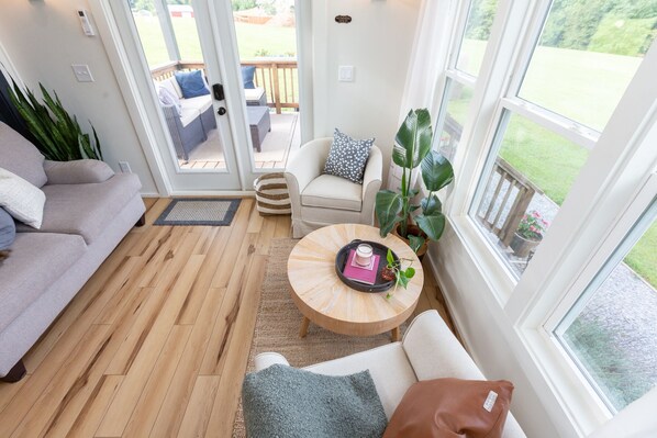 Una sala de estar acogedora con sofá, sillón y mesa de centro. Un ventanal corredizo da a un patio exterior con mesa y sillas.