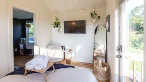 Un dormitorio moderno con cama, escritorio, silla y televisor. Hay plantas y una ventana con vistas.