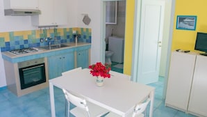 A compact kitchen with a gas stove, sink, and a small oven. There is a dining table with chairs and a vase of flowers. A bathroom is visible through an open door. - Il Carrubo bright and fresh apartment with beautiful view (Marina di Novaglie)