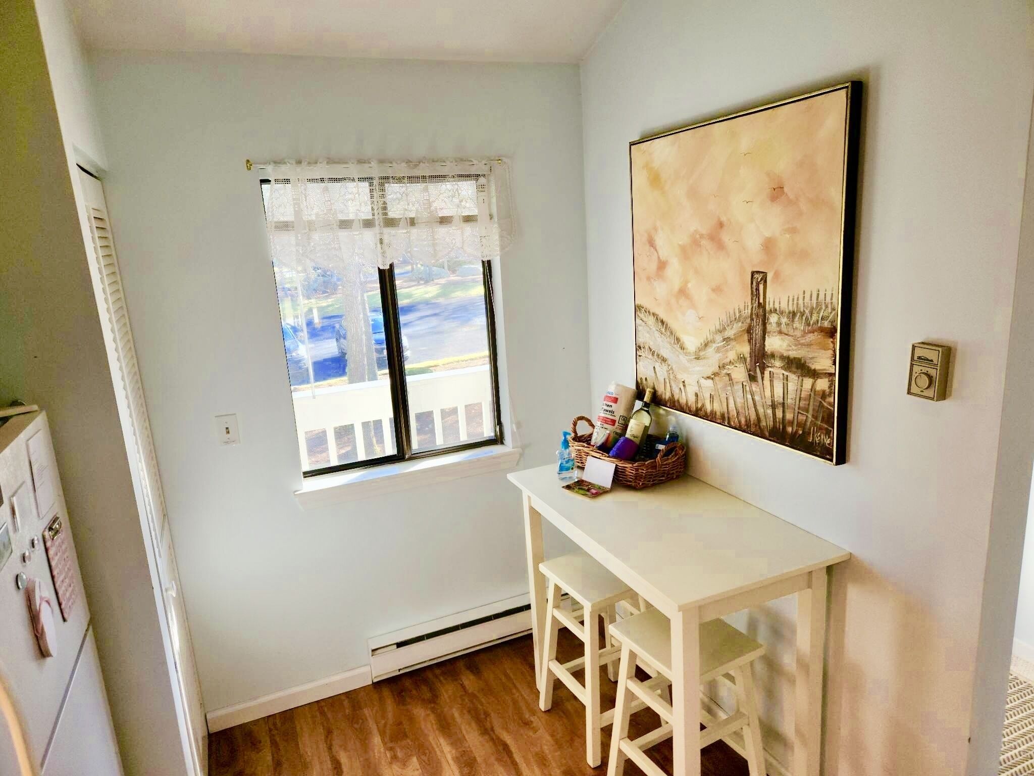A small room with a window, a white table with two chairs, a painting on the wall, and a thermostat.