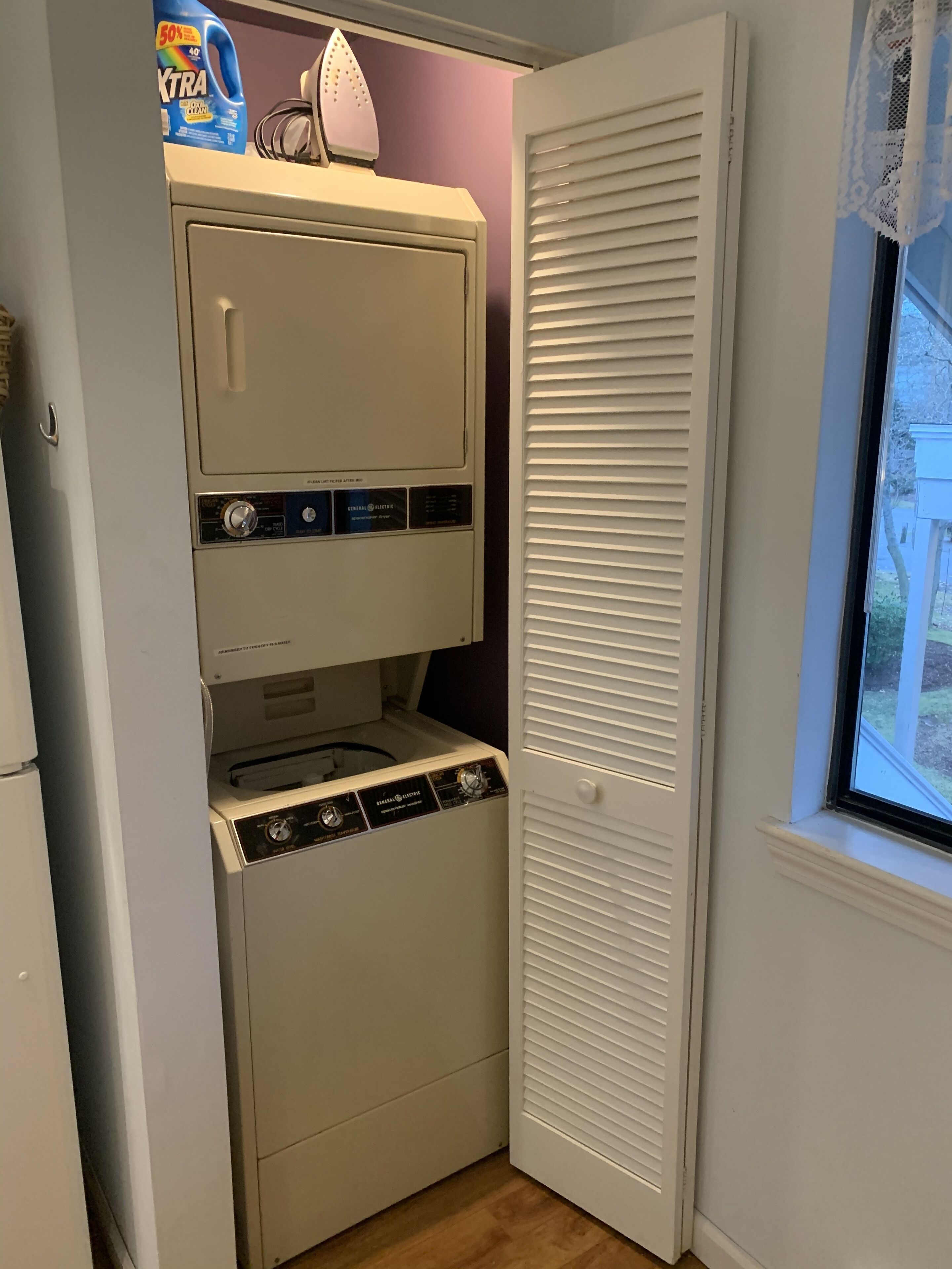 A compact laundry room with a washer and dryer, a ironing board, and a window with blinds.