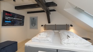 Un dormitorio moderno con techo inclinado, una cama grande con toallas blancas ordenadas, un televisor de pared mostrando un anuncio de Netflix y una obra enmarcada sobre la cama.
