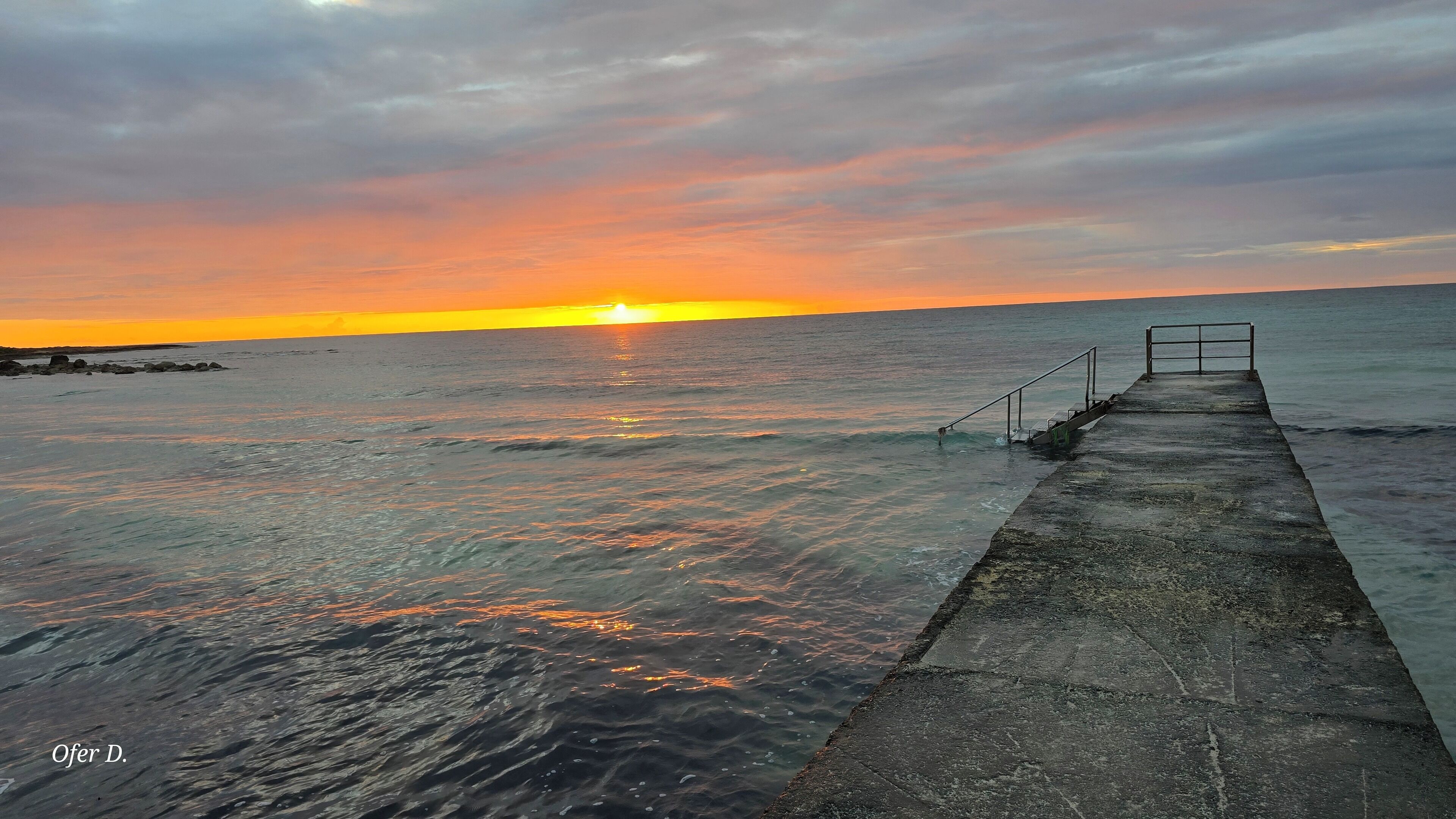 A concrete pier extending into calm waters with a sunset in the background.