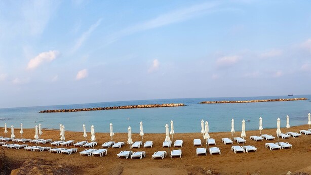 A beach with rows of white sun loungers and umbrellas, a rocky shoreline, and calm sea. - Dahan Sirena lighthouse luxury apartment (Paphos)