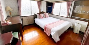 A bedroom with a wooden floor, a bed with white and pink bedding, a bedside table with a lamp, and a window with curtains. - Warayunso Villa (Naha)