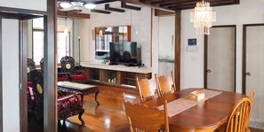 A dining area with a wooden table and chairs, a television on a stand, and a chandelier. - Warayunso Villa (Naha)