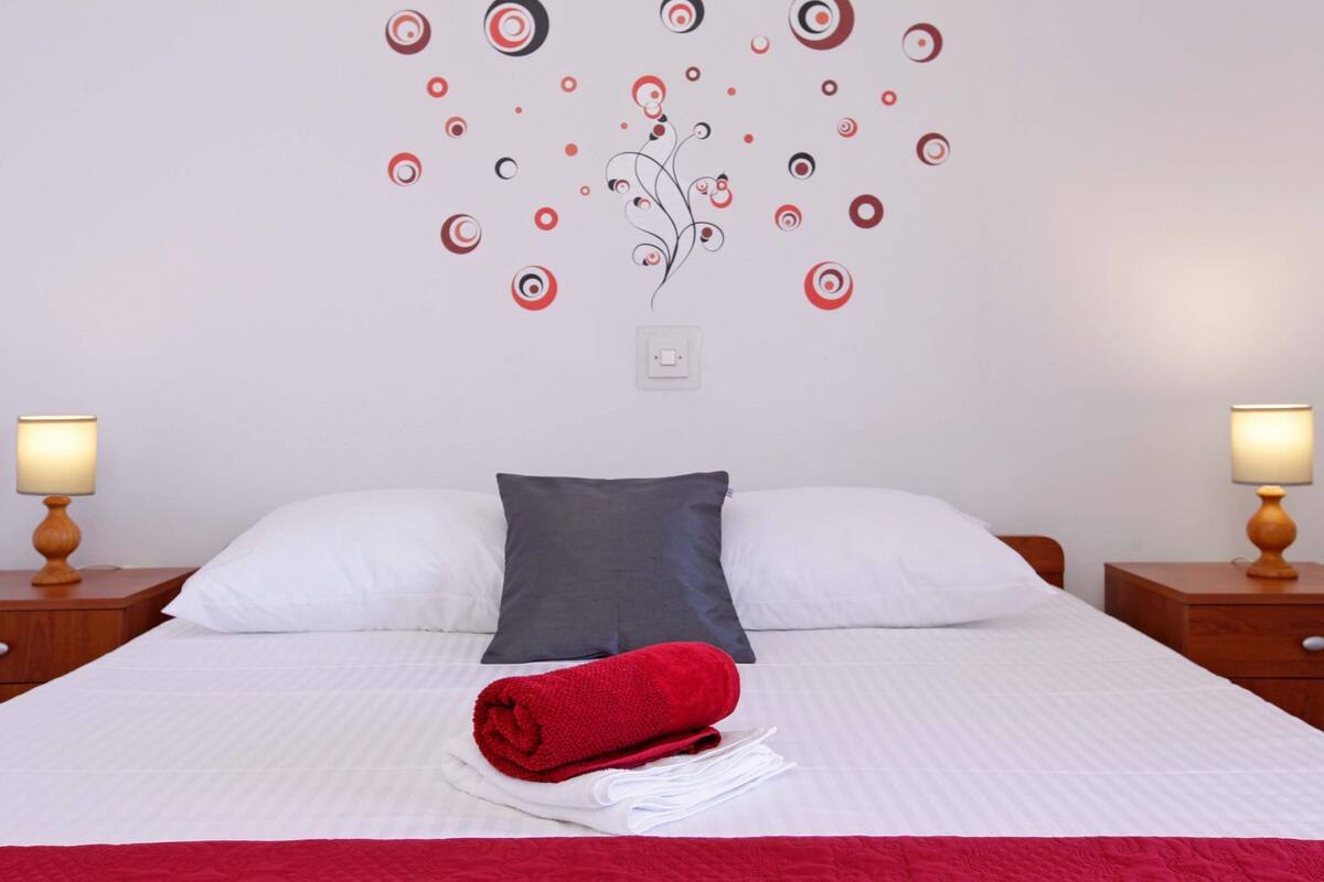 Una cama bien hecha con ropa de cama blanca y roja, dos lámparas de noche y una decoración de pared con diseños circulares.